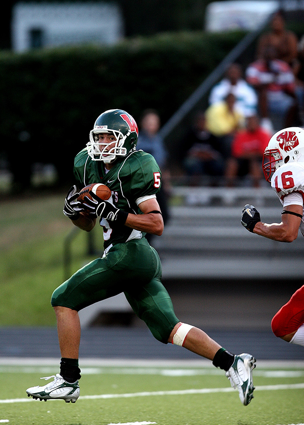 Football player in green running with ball in hands and looking toward camera. Chased by football player in red and white, whose helmet, arm and knee are in frame. 