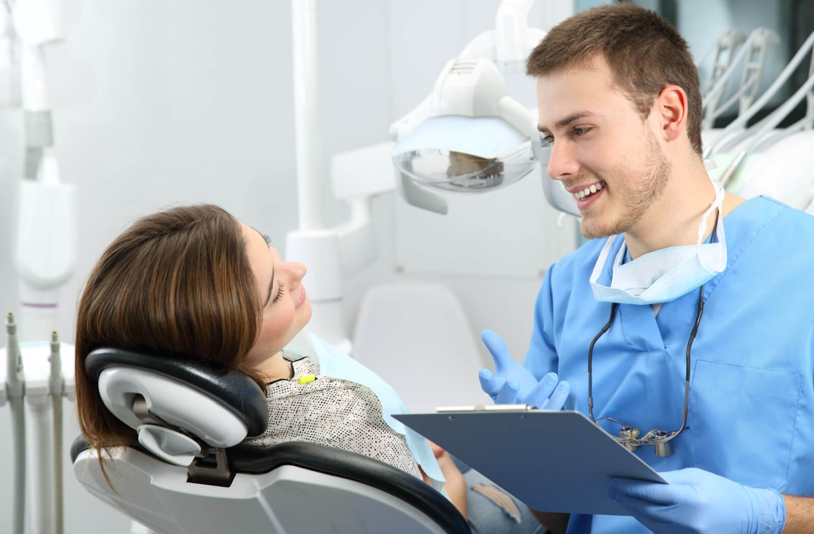 A male dentist in blue scrubs holding a clipboard and smiling at his patient in the dental chair