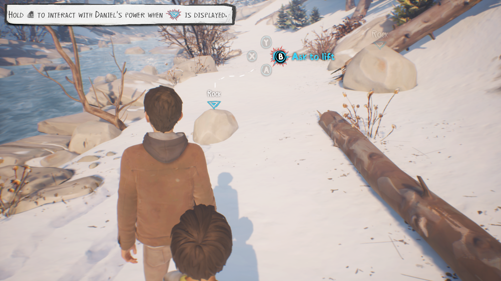 A teenage boy and a smaller boy stand in the snow. In the background is a rock, labelled "Rock" in white writing, with a broken white line leading to a prompt surrounded by blue and pink spikes to press "B" to "Ask to lift". The font is also in bright blue letters. The white writing and label is very difficult to see against the snow. In the upper left corner is prompt that says: "Hold LT to interact with Daniel's power when (the blue and pink spiky icon) is displayed."