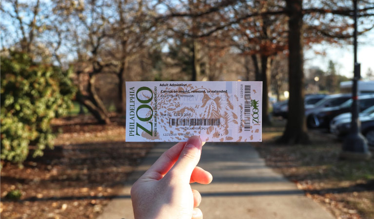 A hand holding up a ticket to the Philadelphia Zoo with an autumn scene in the background.
