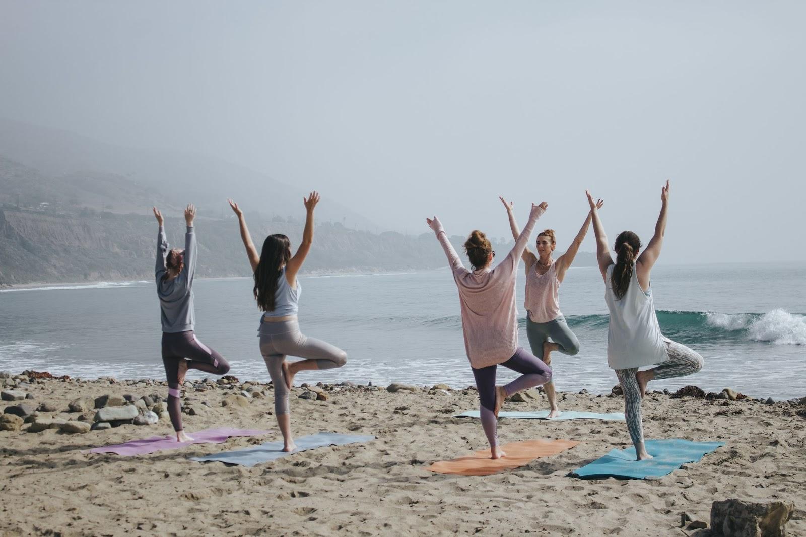 A yoga class commences at a beach. There are a handful of people taking the class. Everyone is wearing pastel colors. 
