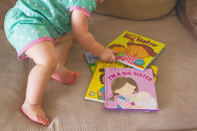 Toddler grabbing a book about being a big sister from a stack of books next to her on a couch.