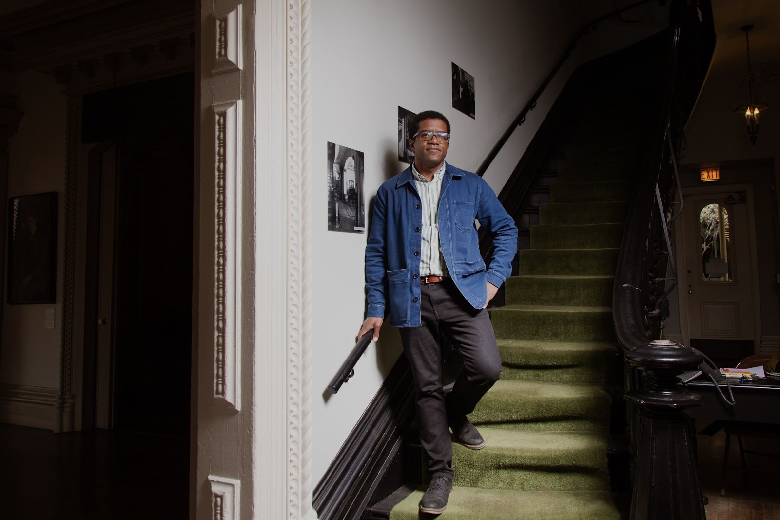 Image: Ross Stanton Jordan stands on the bottom stairs of a dark brown and bright green staircase at the Jane Addams Hull House Museum. On the wall next to hum are historic images. In the background you can see into other unlit parts of the building, with an exit sign glowing above the door at the end of the right corridor. Photo by Joshua Clay Johnson.