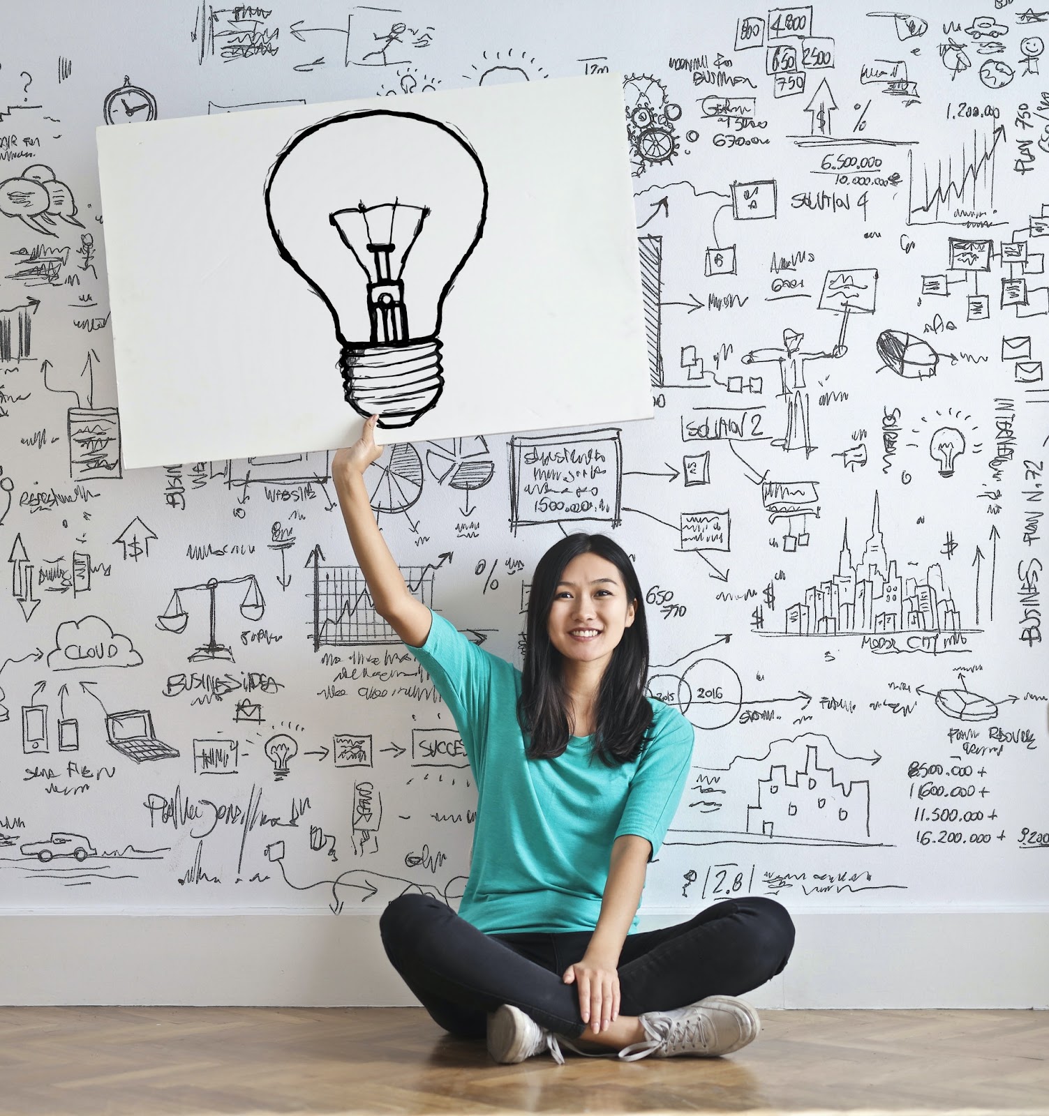 A smiling young woman holds a large poster with a light bulb on it. She is seated in front of a crowded vision board covered in ideas, topics, and how they all relate. Arrows, sketches, and words in black are visible on a white marker board.