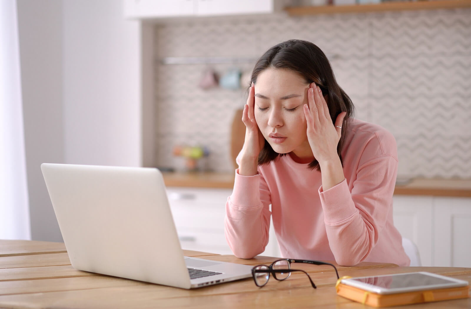 A woman that is sitting in front of her computer for long hours is experiencing eye strain and headaches, she is massaging her temper in both hands while her eyes are closed.
