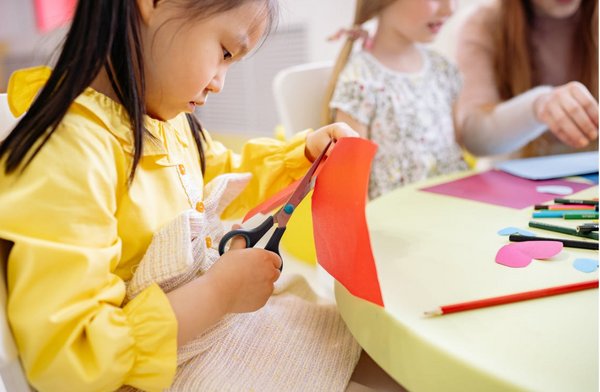 A young children dressed in yellow sits at a table using a pair of black scissors to cut a straight line in an orange piece of paper.