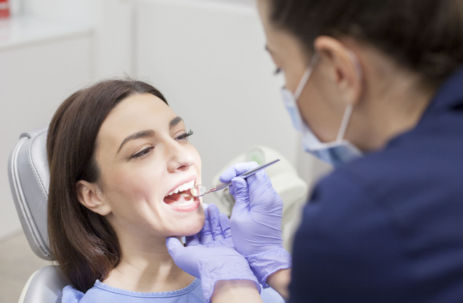 A woman sitting in a dentist's chair smiling while her dentist examines her teeth.
