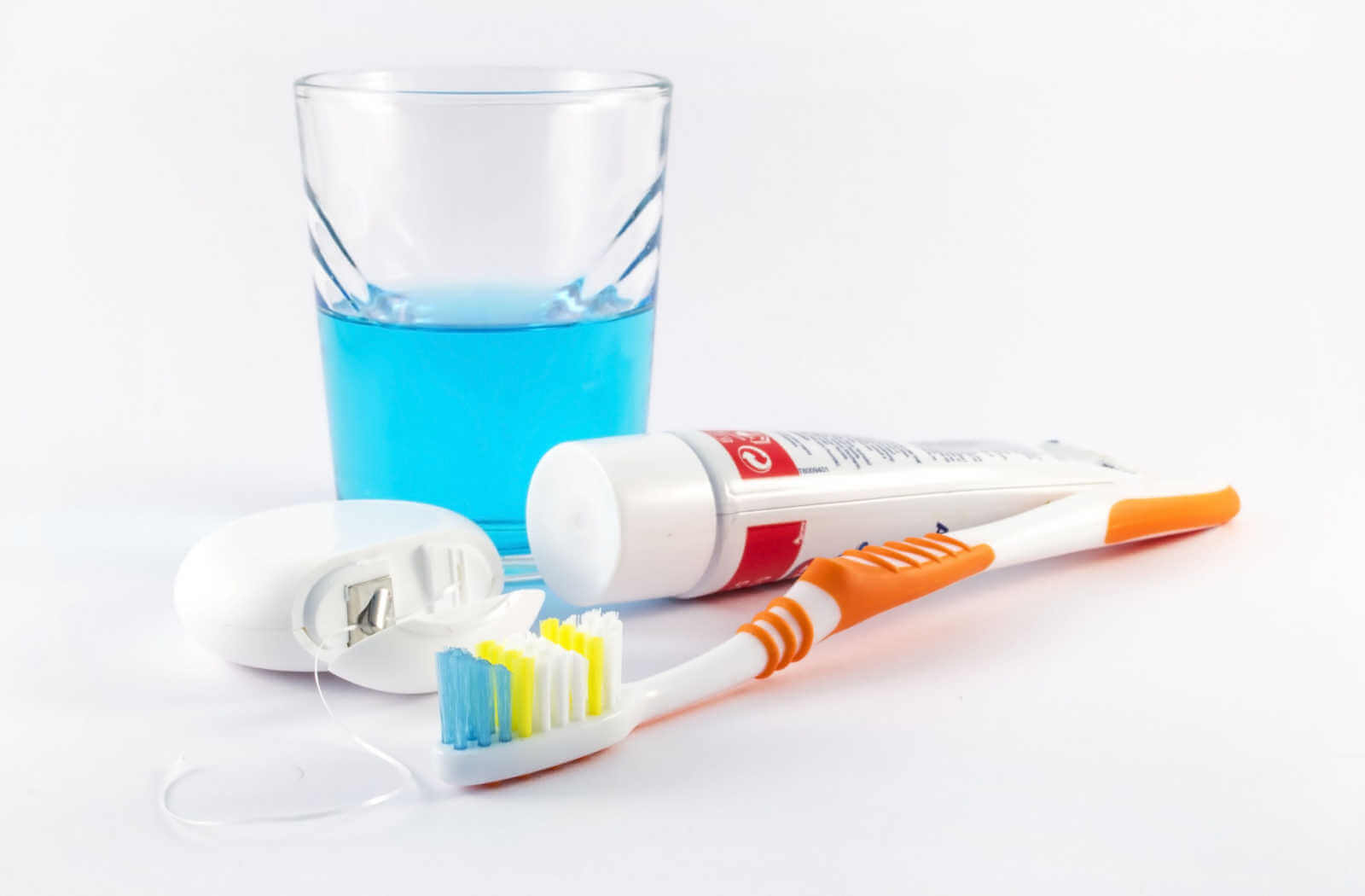 Showing in the photos are of a half glass of blue-coloured mouthwash, toothpaste in a white tube, a  small case of the dental floor, and an orange-coloured toothbrush.