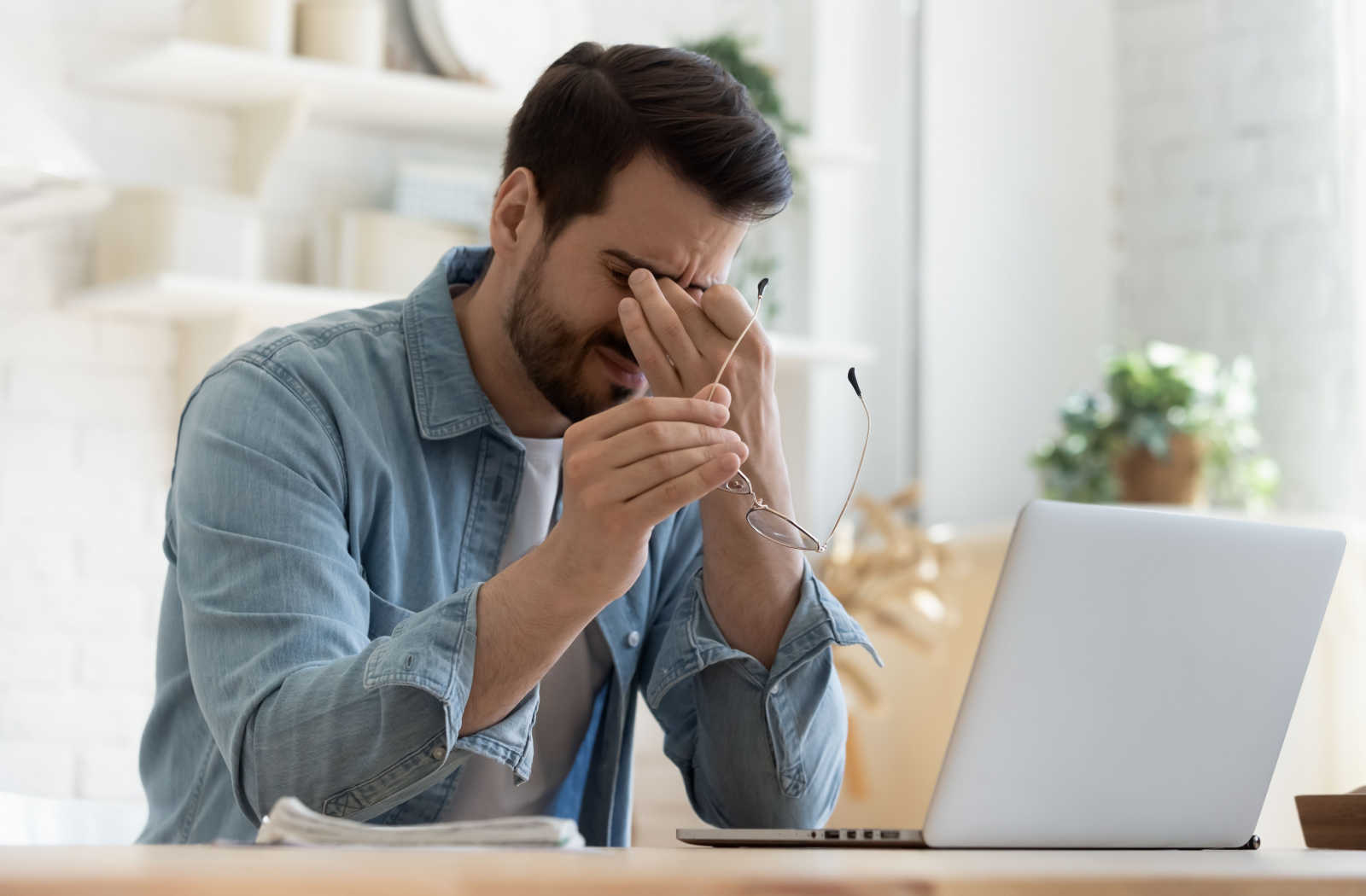 A man holding a pair of glasses in one hand and squinting his eyes while experiencing digital eye strain due to constantly working on a laptop.