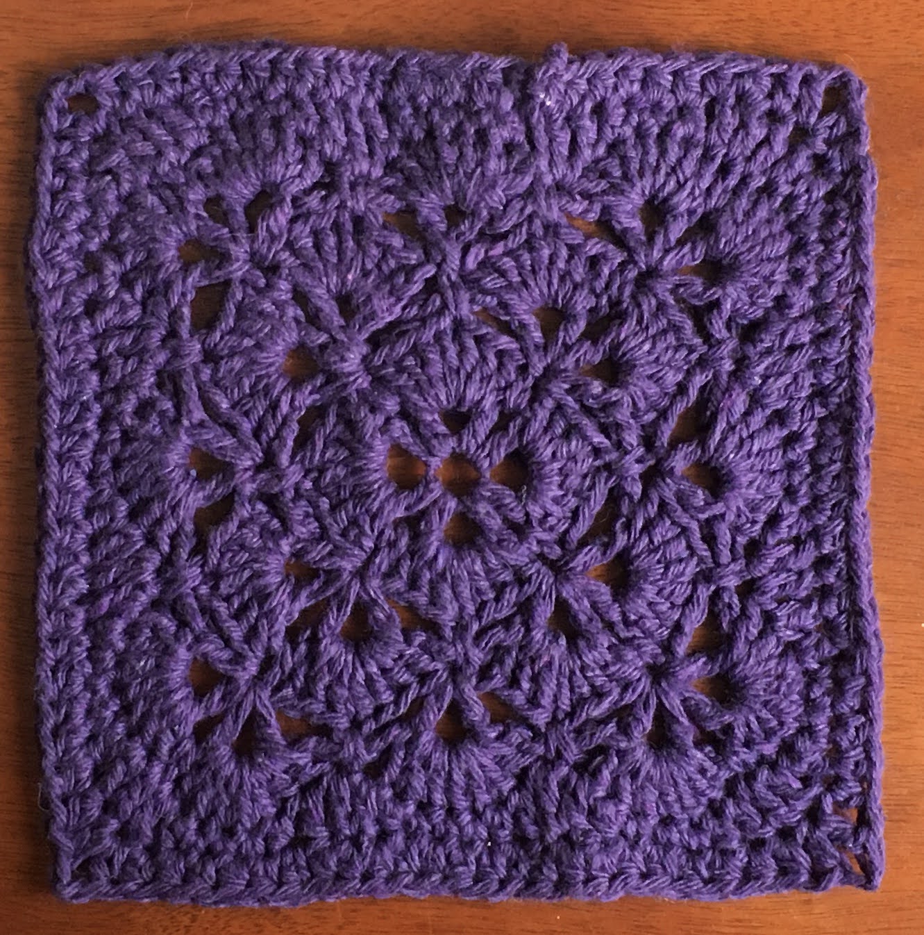 A deep purple crocheted square with a floral, fan-like design.