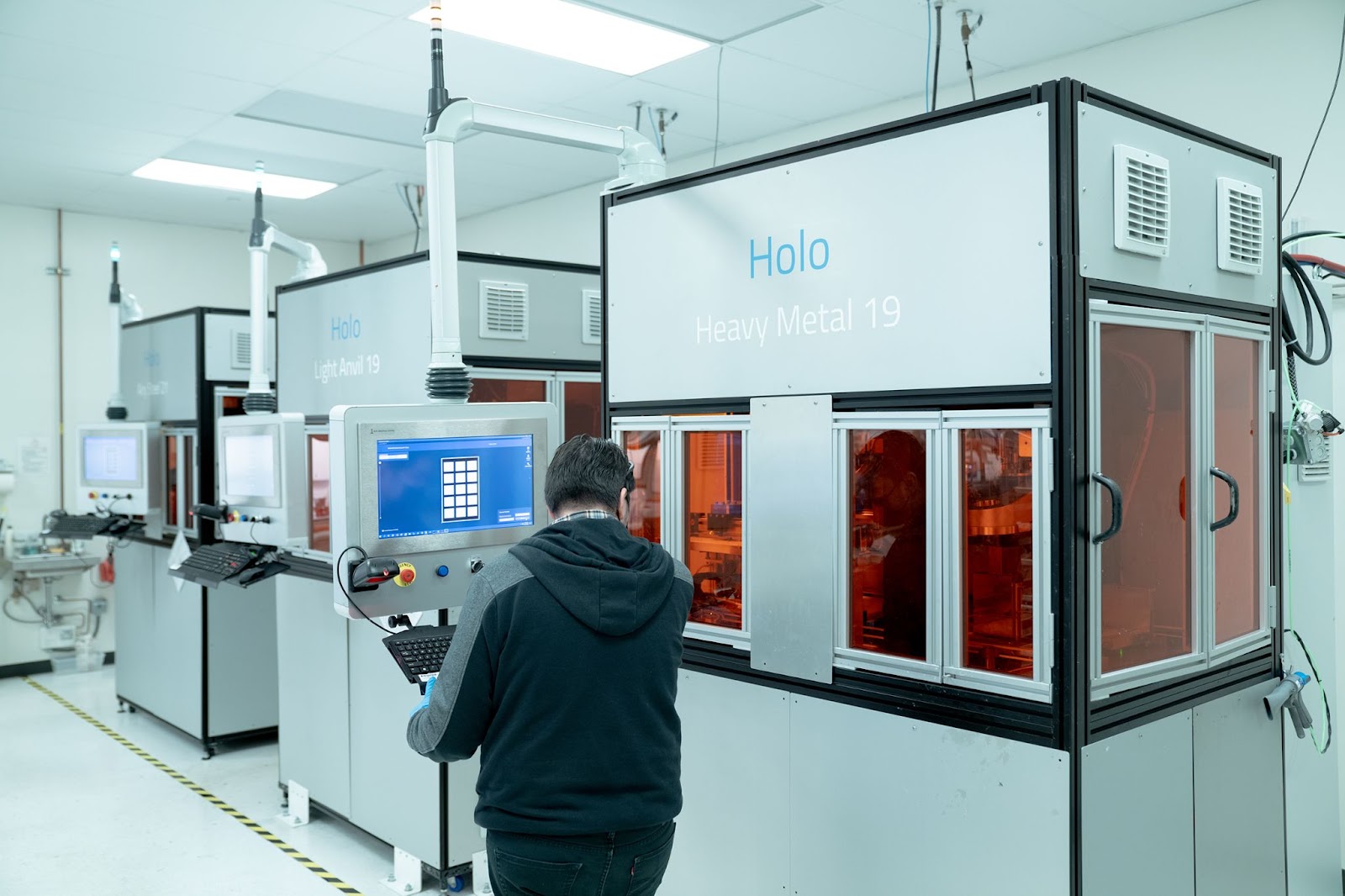 A 3D printer operator interacts with the control screen on one of Holo's PureForm™ metal 3D printers, with two more printers from the Holo fleet in the background