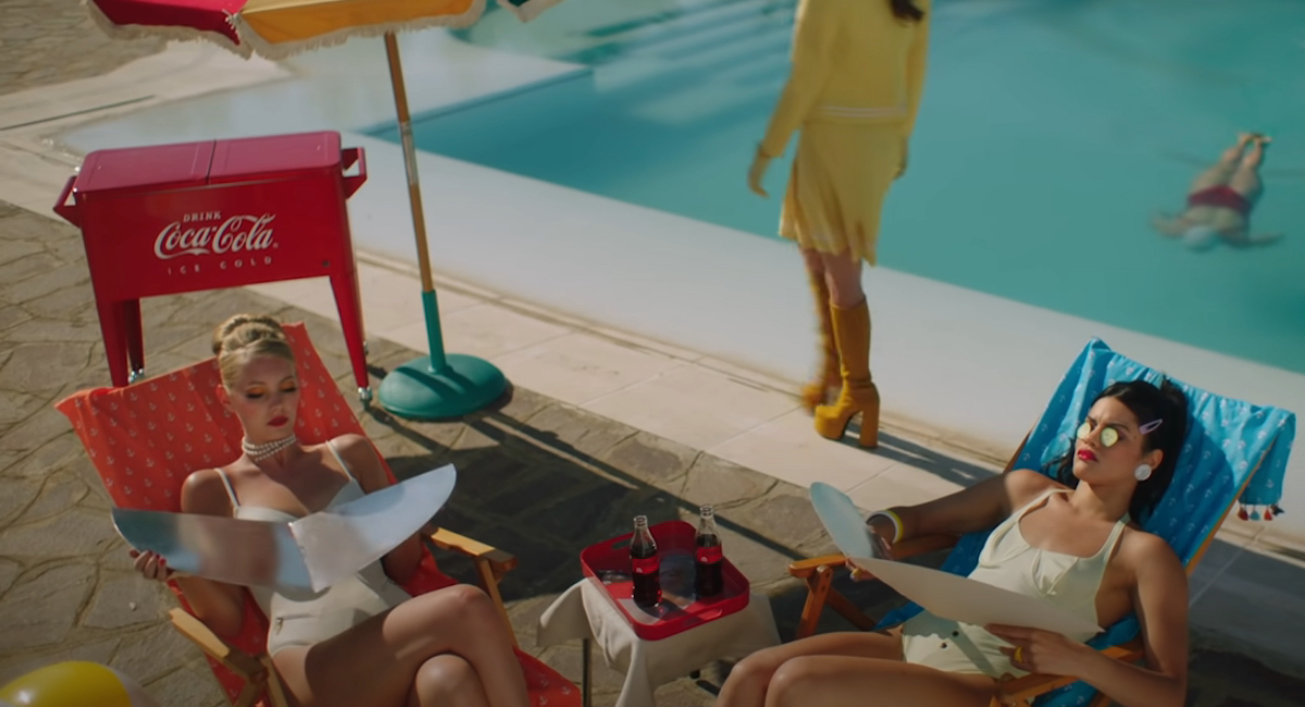 Scena dal video "Mille", la nuova hit estiva di Fedez, Orietta Berti e Achille Lauro. La scena mostra due ragazze che prendono il sole e sullo sfondo il logo CocaCola.