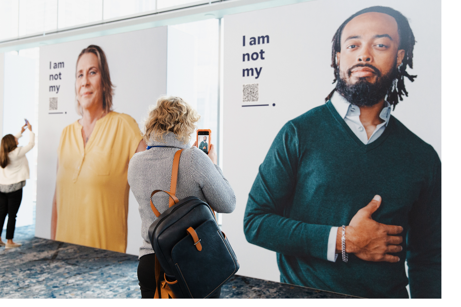 Image shows two women taking photos of the displays at the Futureworks event. Displays feature individuals with the caption "I am not my..." with a QR code to scan to learn more.